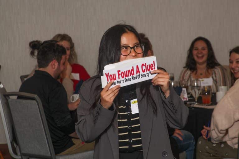 A woman in glasses smiles while holding a sign that reads You Found A Clue!—a great intro text for your front page, set in a lively room where people are seated at tables, laughing and enjoying themselves.