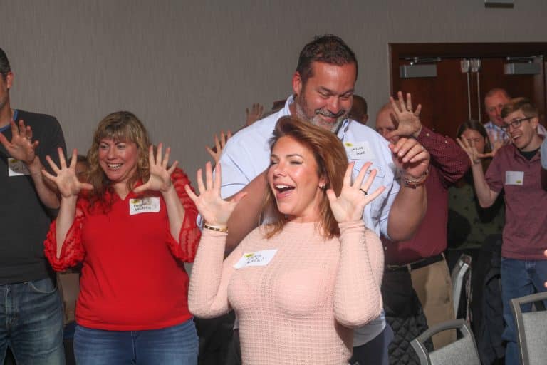 A group of adults stand closely together indoors, smiling and holding their hands up with fingers spread wide, appearing to participate in a fun Front Page group activity or icebreaker.