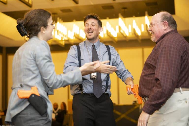 Three adults—two men and one woman—in business attire stand in a well-lit room laughing together. With orange toy guns holstered at their sides, this scene could be the perfect Front Page intro text for a playful corporate training session.