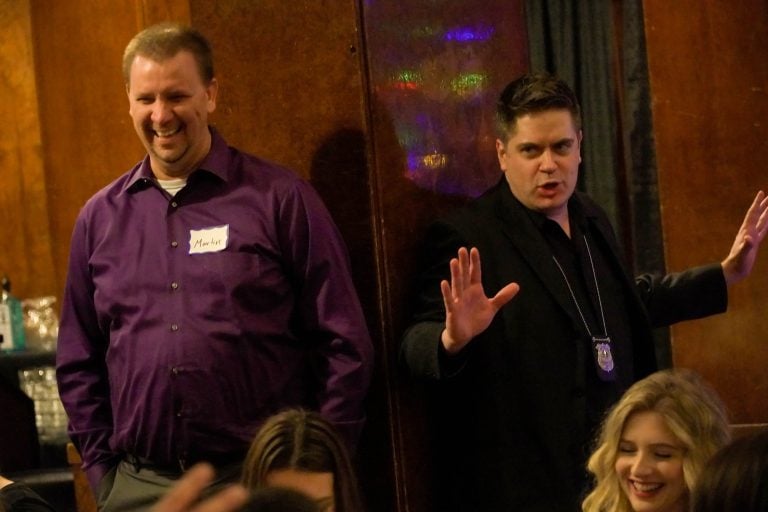 Two men stand indoors; one on the left, smiling with a name tag reading Mark, and the other on the right, gesturing with his hands and wearing a badge around his neck. A woman with blonde hair sits in the foreground, perfect for Intro Text or Front Page.