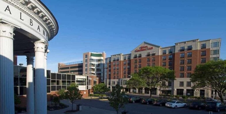 A view of a city street with a round white building featuring columns on the left and a large brick hotel with Hilton Garden Inn signage on the right, trees and parked cars in the foreground under a clear blue sky.