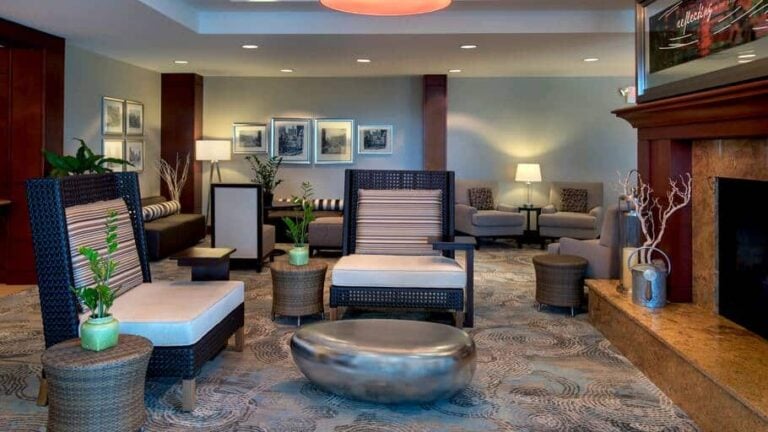 A modern hotel lobby features wicker chairs, small tables with plants, a metallic oval ottoman, neutral-toned sofas, framed artwork on the walls, and soft lighting creating a welcoming atmosphere.