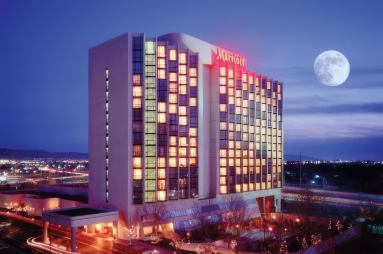 A tall Marriott hotel building is illuminated at night, with many windows lit up. The full moon shines brightly in a clear evening sky, and city lights are visible in the background.