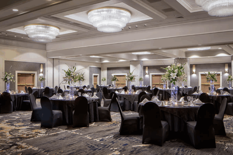 A spacious banquet hall set for an event, featuring round tables with black chairs, formal place settings, and tall floral centerpieces, under large, elegant chandeliers.