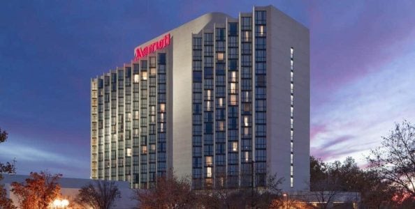 A tall hotel building with the Marriott logo illuminated at the top. The structure features many windows and is set against a twilight sky with scattered clouds. Trees with autumn foliage are visible in the foreground.