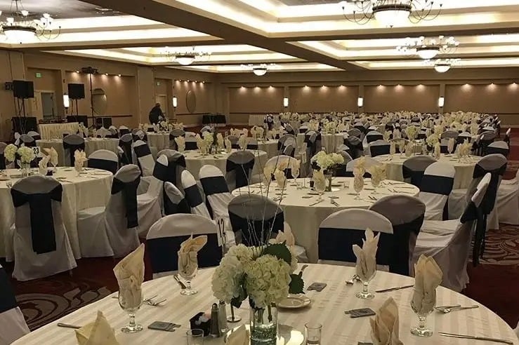 A large banquet hall set up for an event, featuring round tables with white tablecloths, dark chair sashes, neatly folded napkins, glassware, and white floral centerpieces, under warm overhead lighting.