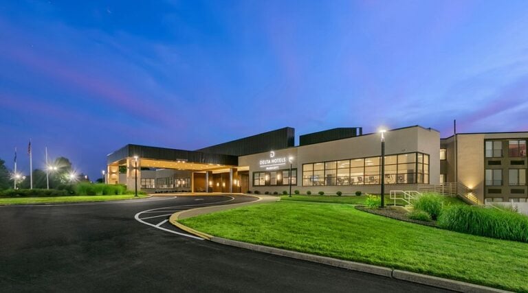 A modern hotel building with large windows and a covered entrance, illuminated by exterior lights at dusk. The sky is a gradient of blue and purple, and the landscaping features green grass and shrubs.