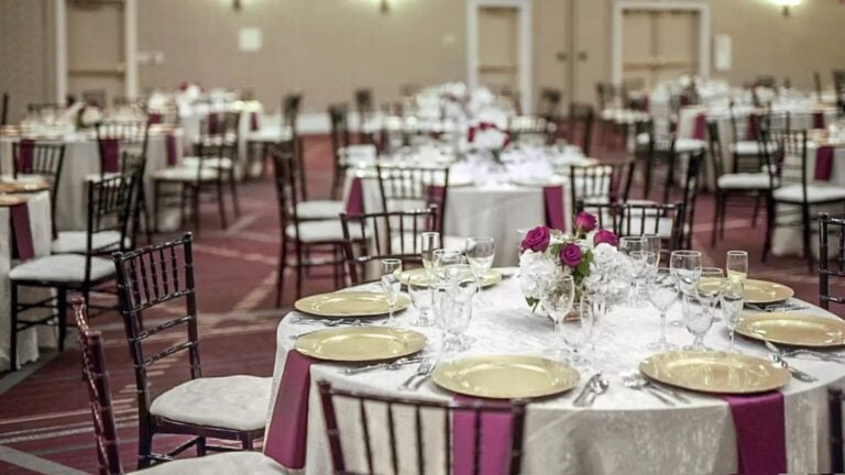 A banquet hall with round tables set for an event. Each table has gold plates, glassware, napkins, and a floral centerpiece with pink and white flowers. Dark wooden chairs surround the tables on a carpeted floor.