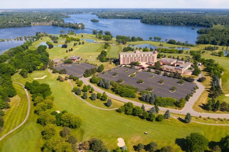 Aerial view of a large hotel and parking lot surrounded by a golf course with sand traps, lush green trees, and several lakes, with forests and more water in the background under a blue sky.
