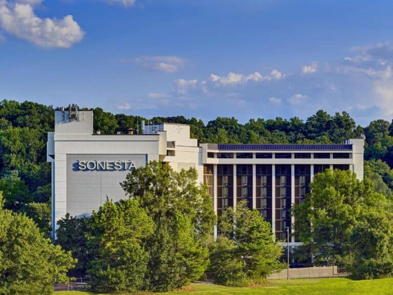 A multi-story Sonesta hotel building is partially obscured by tall green trees, set against a backdrop of dense forest and a blue sky with scattered clouds.