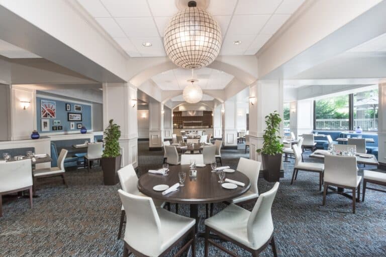 Spacious, modern restaurant with white chairs, dark round tables set for dining, large windows letting in natural light, hanging globe lights, potted plants, and framed art on blue accent wall.