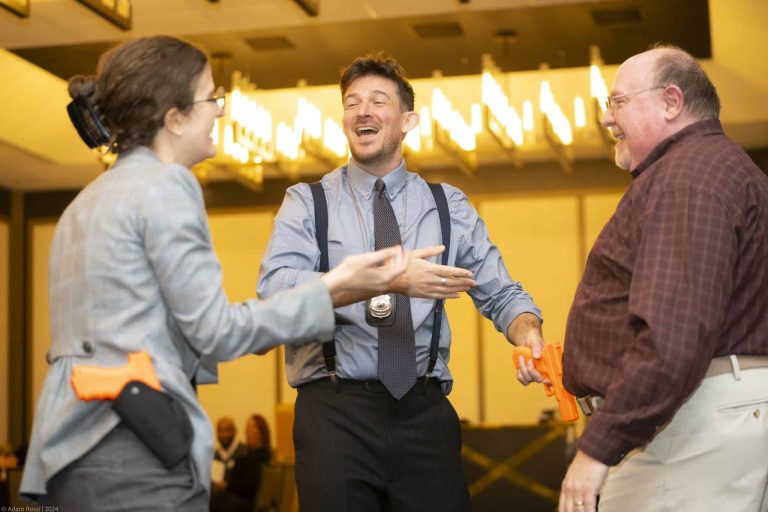 Three adults indoors, two holding orange toy guns, laugh and interact in a well-lit room. One man wears a badge and suspenders, suggesting a police role-play or training exercise. People are seated in the background.