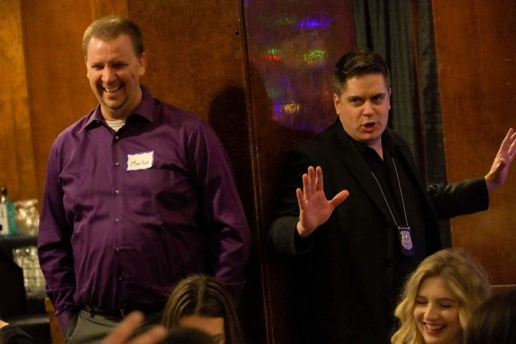 Two men stand indoors; one, wearing a purple shirt and name tag, laughs, while the other, in a dark jacket with a badge necklace, gestures with hands raised. Two women sit in the foreground.