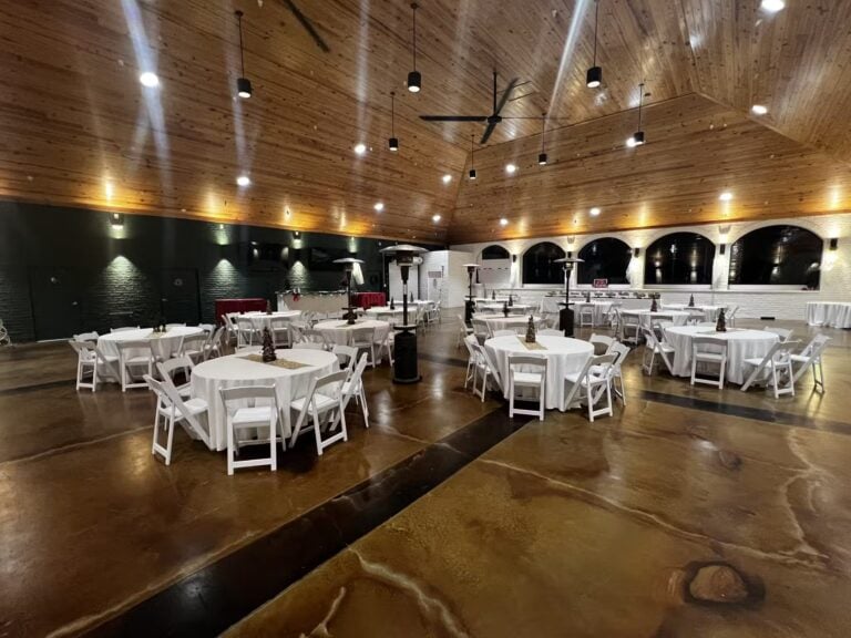 A spacious event hall with a high wooden ceiling, polished brown floors, and white round tables with matching chairs arranged for a gathering. The room is well-lit and has a modern, rustic design.