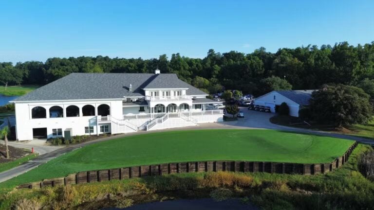 A large white clubhouse building with a gray roof overlooks a manicured green lawn, surrounded by trees and a blue sky, with a golf cart path and a separate barn-like structure nearby.