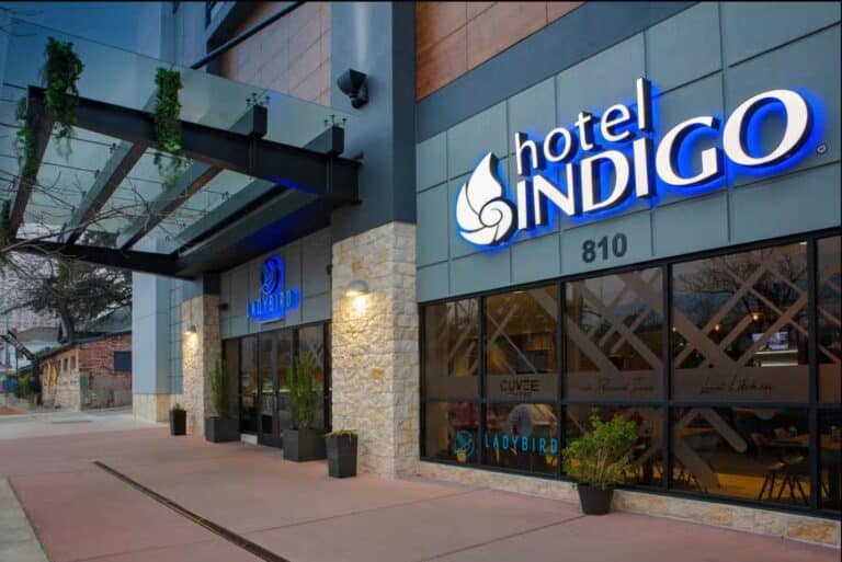 The entrance of Hotel Indigo with modern glass and stone facade, large blue and white illuminated sign, potted plants, and address number 810 visible on the building.