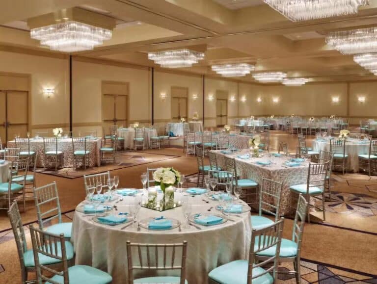 A banquet hall set for an event with round tables covered in light blue tablecloths, matching napkins, silver chairs, and elegant floral centerpieces. Chandeliers hang from the ceiling, creating a bright, formal atmosphere.