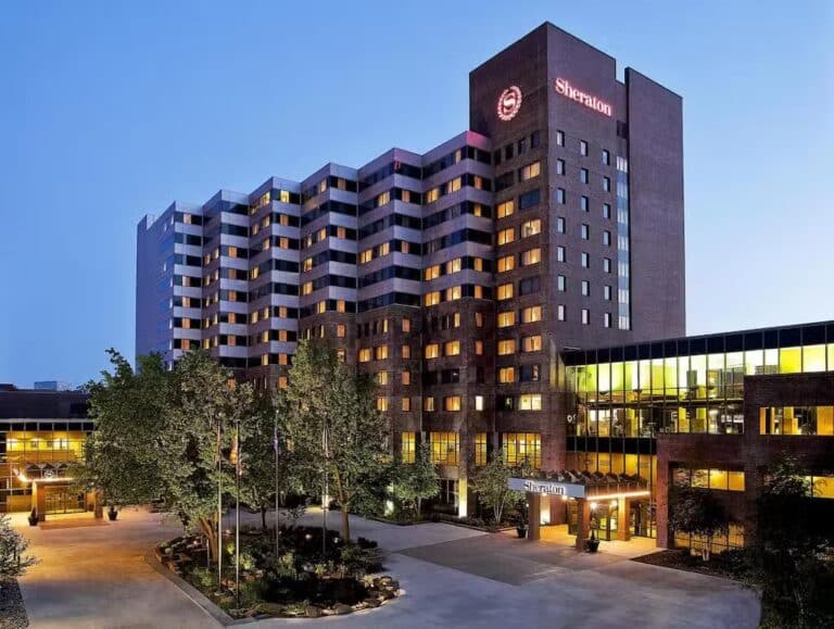 A multi-story Sheraton hotel building is illuminated at dusk, with many windows lit. Trees and a circular driveway are in front of the main entrance, and the Sheraton logo is visible on the building.