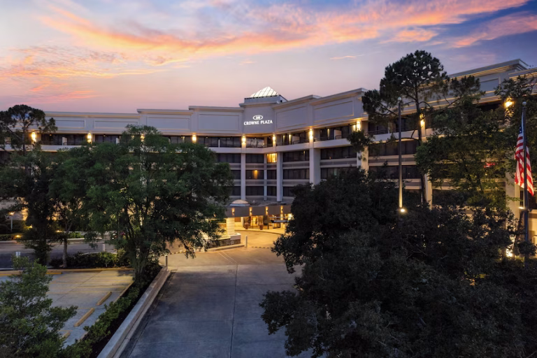 A multi-story hotel building with “Crowne Plaza” signage, surrounded by trees and a parking lot, is shown at sunset with a colorful sky in the background. An American flag is visible on the right.