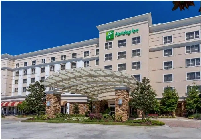 A Holiday Inn hotel with a modern beige exterior, large windows, and a covered driveway entrance supported by stone pillars, surrounded by greenery under a clear blue sky.