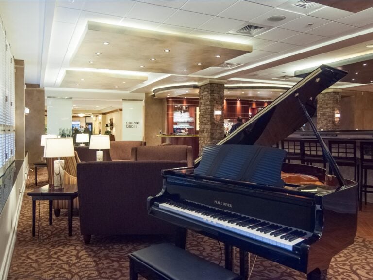 A grand piano sits in a warmly lit hotel lounge with brown sofas, lamps, and stone pillars. The room features overhead lighting, carpeted floors, and a bar area in the background.
