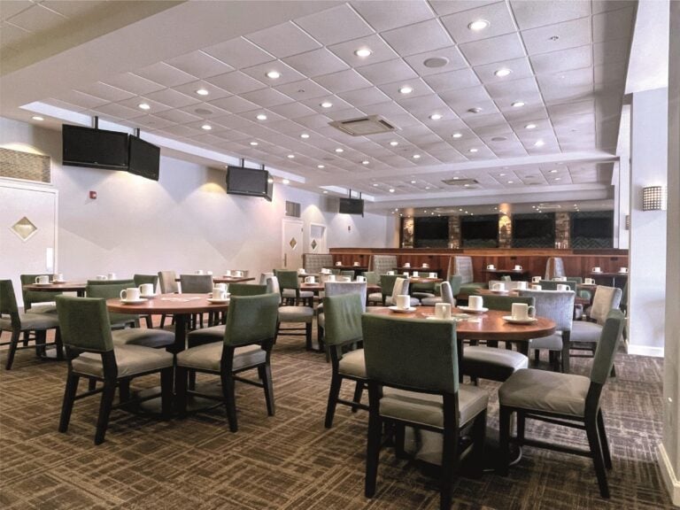 A spacious dining area with several round tables and cushioned chairs, set with white mugs. Overhead lights brighten the room, and TV screens are mounted on the walls. The atmosphere is modern and clean.