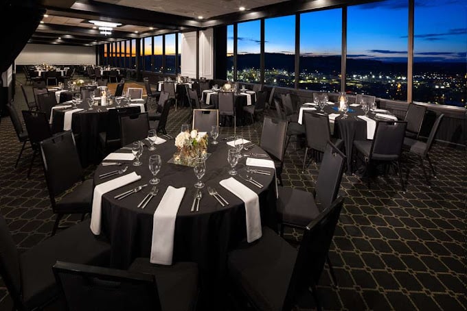 Elegant banquet hall set for an event with round tables covered in black tablecloths, white napkins, glassware, and floral centerpieces. Large windows reveal a cityscape at sunset in the background.