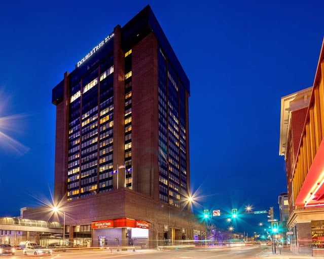 Tall brick DoubleTree hotel building illuminated at night, located on a busy city street corner with traffic lights and light trails from passing cars. The sky is dark blue, indicating early evening.