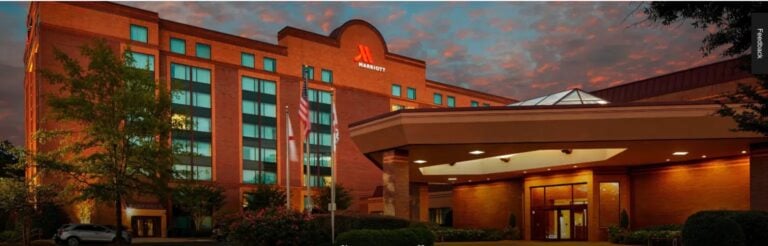 A large brick Marriott hotel building at dusk, with glowing lights inside, an entrance canopy, an American flag, trees, and a dramatic, colorful sky in the background.