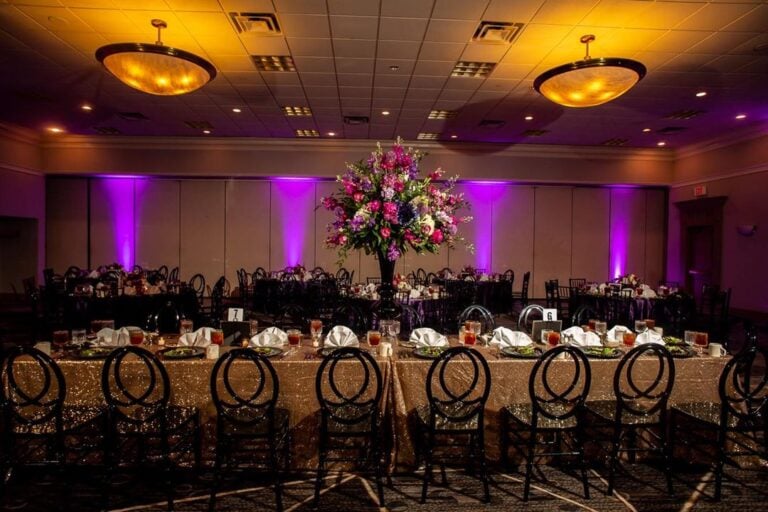 A banquet hall with round tables, a long table with gold sequin cloth, black chairs, neatly set plates and drinks, and a large floral centerpiece; purple uplighting creates a festive atmosphere.