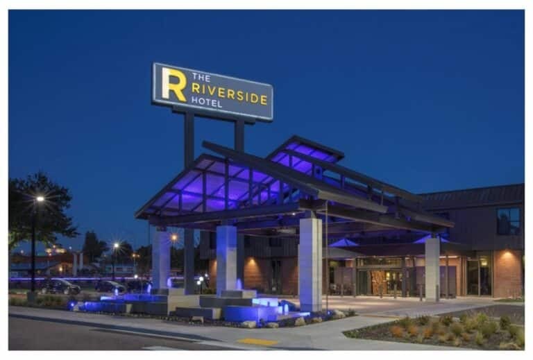 The exterior of The Riverside Hotel at night, featuring a modern entrance with purple-lit architectural details and a large illuminated sign above the driveway.