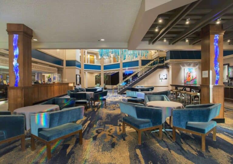 Modern hotel lobby with blue velvet chairs and sofas, round tables, patterned carpet, illuminated columns, a bar on the left, and a staircase leading to an upper level with hanging blue decorations.