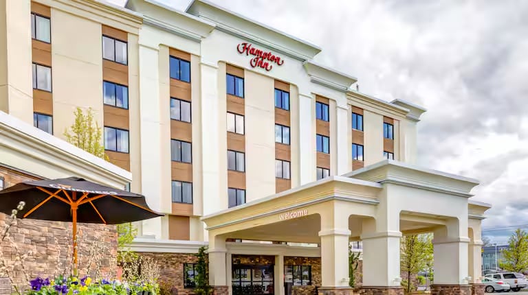 A multi-story Hampton Inn hotel with a tan and white exterior, stone accents, large windows, and a covered entrance. An umbrella and plants are visible in the foreground under a cloudy sky.