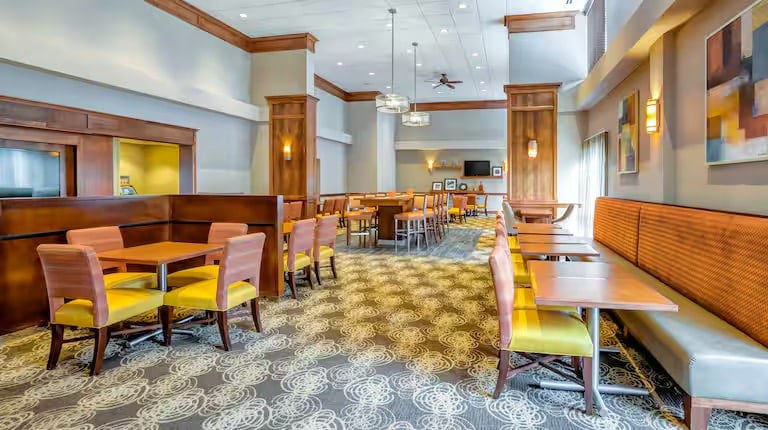 Bright, modern dining area with wooden tables, yellow cushioned chairs, patterned carpet, and large windows. Pendant lights hang from a high ceiling, and abstract art decorates the walls. Room is empty of people.