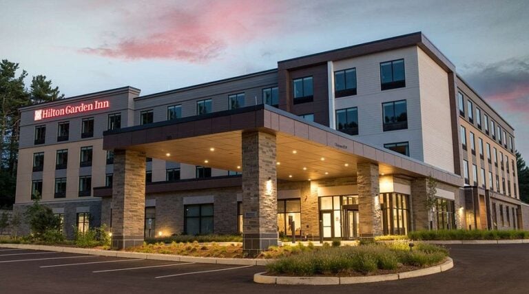 The image shows the exterior of a modern Hilton Garden Inn hotel at sunset, featuring a covered entrance, large windows, and a well-maintained landscape with greenery and a parking area in front.