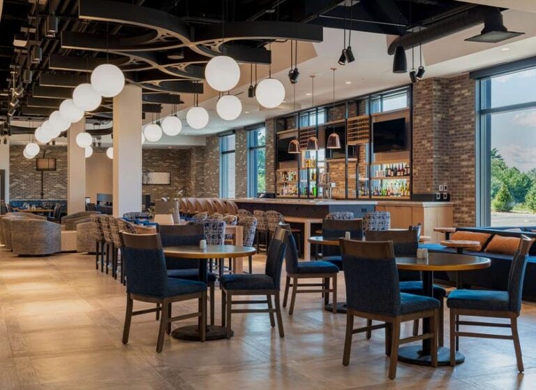 Modern, spacious bar and lounge area with wooden tables and upholstered chairs, globe pendant lights, a brick accent wall, stocked bar shelves, and large windows letting in natural light.