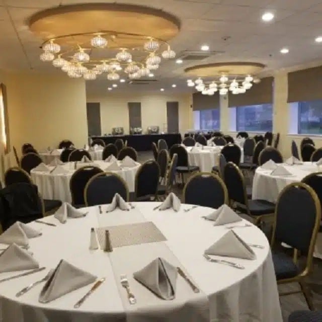 A banquet hall set up for an event, with round tables covered in white tablecloths, neatly arranged place settings, and cushioned chairs. Elegant chandeliers hang from the ceiling, and large windows line one wall.