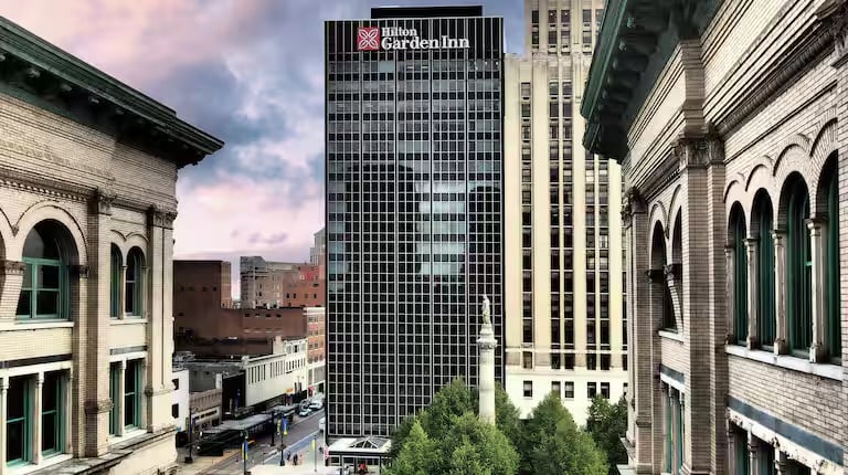 A tall, modern Hilton Garden Inn hotel building stands between two older, ornate buildings with green window frames in an urban area, with a few trees and a street visible below at sunset.