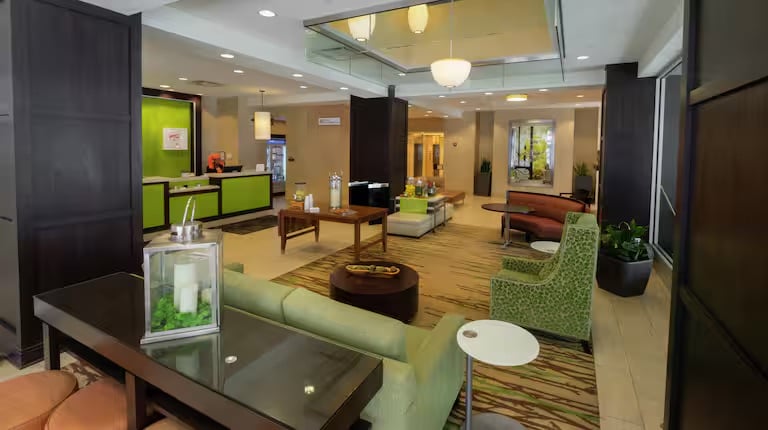 Modern hotel lobby with green and brown decor, comfortable seating areas, a reception desk, coffee table, plants, and contemporary lighting. The space is clean, bright, and inviting with a welcoming ambiance.