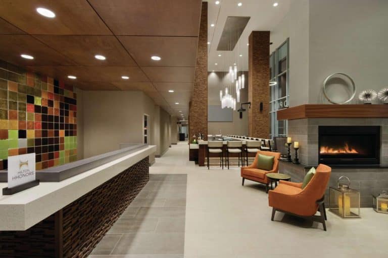 Modern hotel lobby with tiled reception desk, colorful wall art, and a cozy seating area featuring orange chairs near a lit fireplace. Hanging lights illuminate a wooden ceiling, and a high table with stools stands in the background.