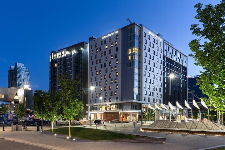 A modern hotel building illuminated at dusk with multiple windows lit up. The foreground features a landscaped area with trees and a small pavement, while adjacent high-rise buildings are visible in the background under a clear sky.