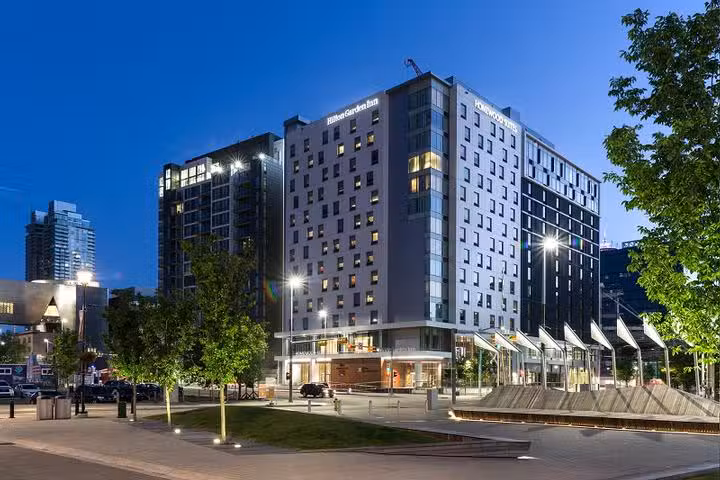 A modern hotel building is illuminated at dusk, surrounded by trees and streetlights, with other high-rise buildings in the background and a clear evening sky overhead.