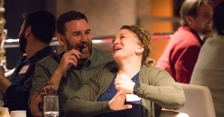 A man and a woman laughing together at a dinner event, with the man playfully holding a prop in his mouth.