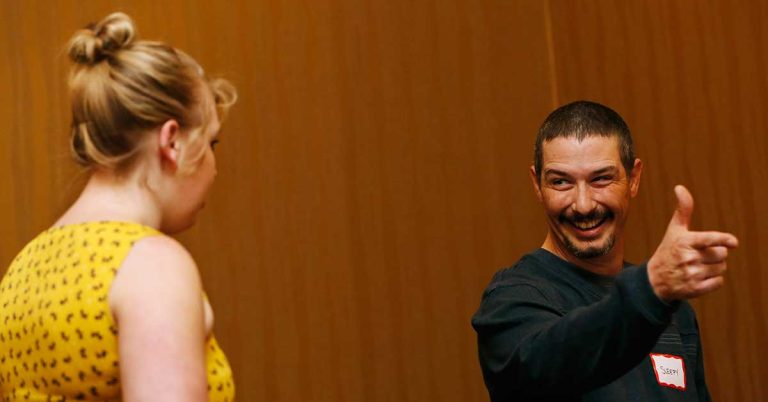 A man playfully pointing finger guns at a woman in a yellow dress while smiling during an interactive event.