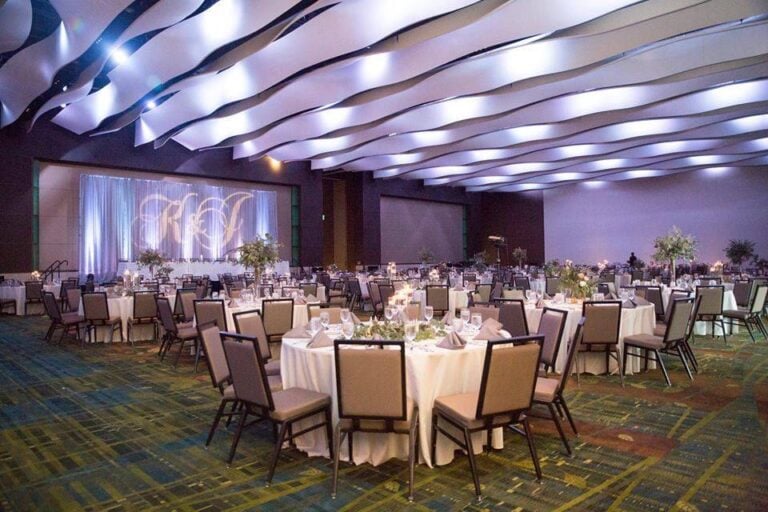 A large, elegant banquet hall with round tables covered in white tablecloths, neatly arranged chairs, soft overhead lighting, and modern wavy ceiling panels, prepared for a formal event or wedding reception.