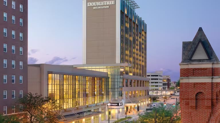 A modern DoubleTree by Hilton hotel building with large glass windows, illuminated in the evening, stands tall in an urban setting with nearby streets and other buildings visible.