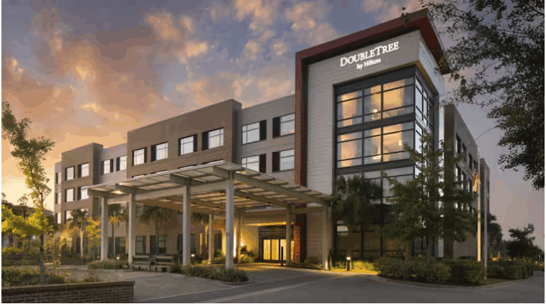 A modern DoubleTree by Hilton hotel building with four stories, large windows, and a covered entrance, surrounded by greenery at sunset.