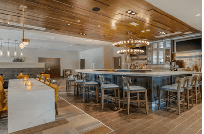 Modern bar area with a marble counter, high chairs, circular light fixtures, and wood-paneled ceiling. The space is well-lit, with a mix of neutral and warm tones, and features a small bar with shelves of bottles.