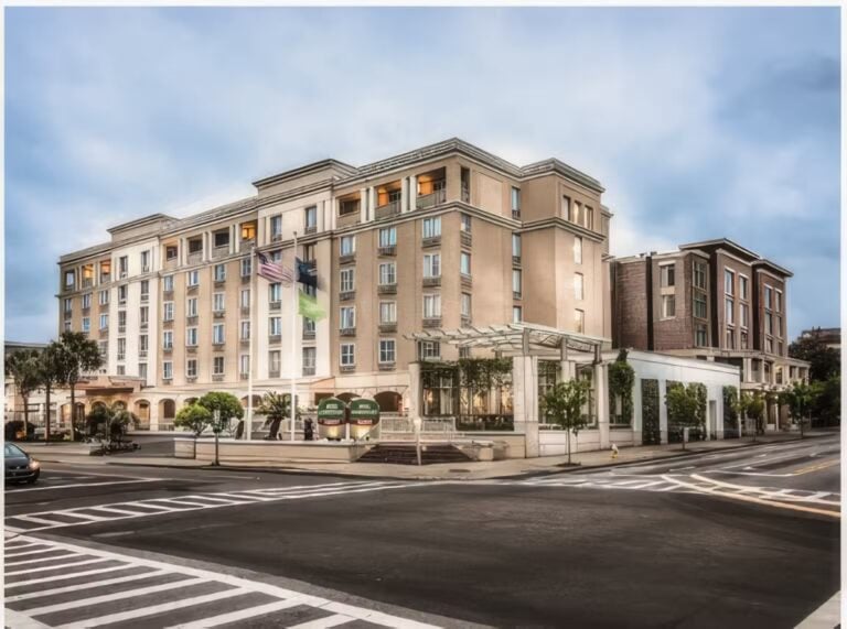 A multi-story beige hotel with large windows and balconies sits at a street corner. The hotel has a glass entrance canopy, American and green flags, and is bordered by trees and crosswalks under a cloudy sky.