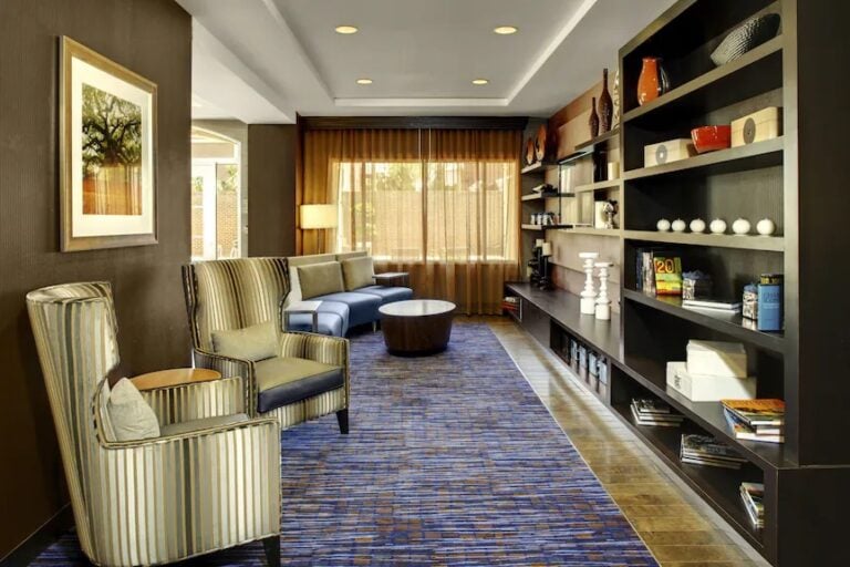Modern lounge with striped armchairs, a sectional sofa, round coffee tables, a blue patterned carpet, wall art, and large built-in shelves filled with books and decorative items, lit by ceiling lights and natural light from large windows.